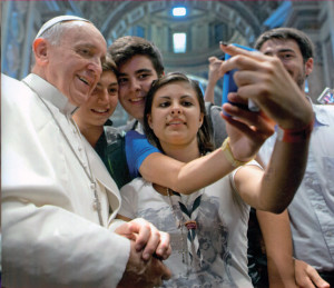 A smiling Pope Francis takes a “selfie” photograph with young people in St. Peter’s Basilica. 